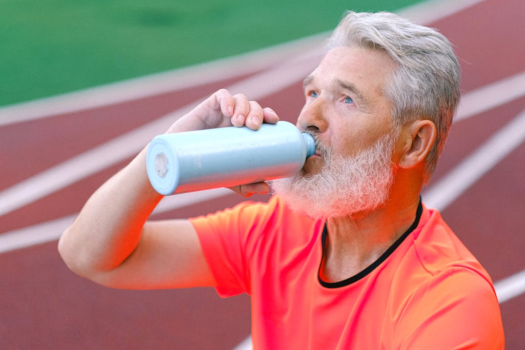 Elderly male with gray beard drinking liquid from thermos while resting after training on blurred background of racetrack